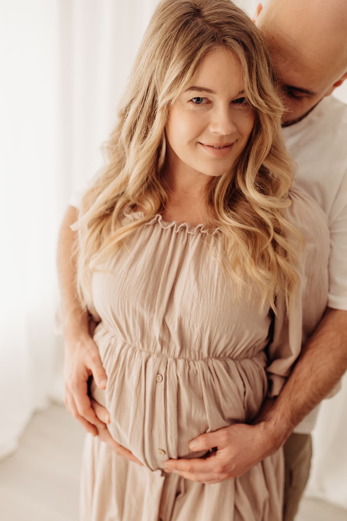 Schwangere Frau schaut lächelnd in die Kamera, ihr Partner hält sie sanft von hinten — Babybauch-Shooting im Tageslichtstudio Bottrop