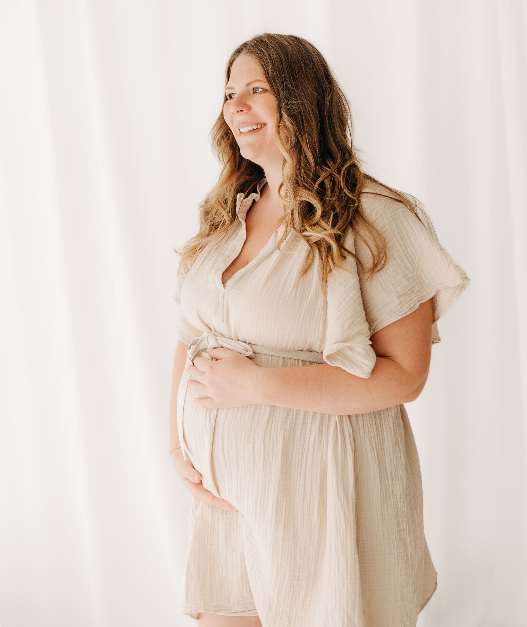 Schwangere Frau im beigen Kleid hält lächelnd ihren Bauch — Babybauch-Shooting im Tageslichtstudio Bottrop