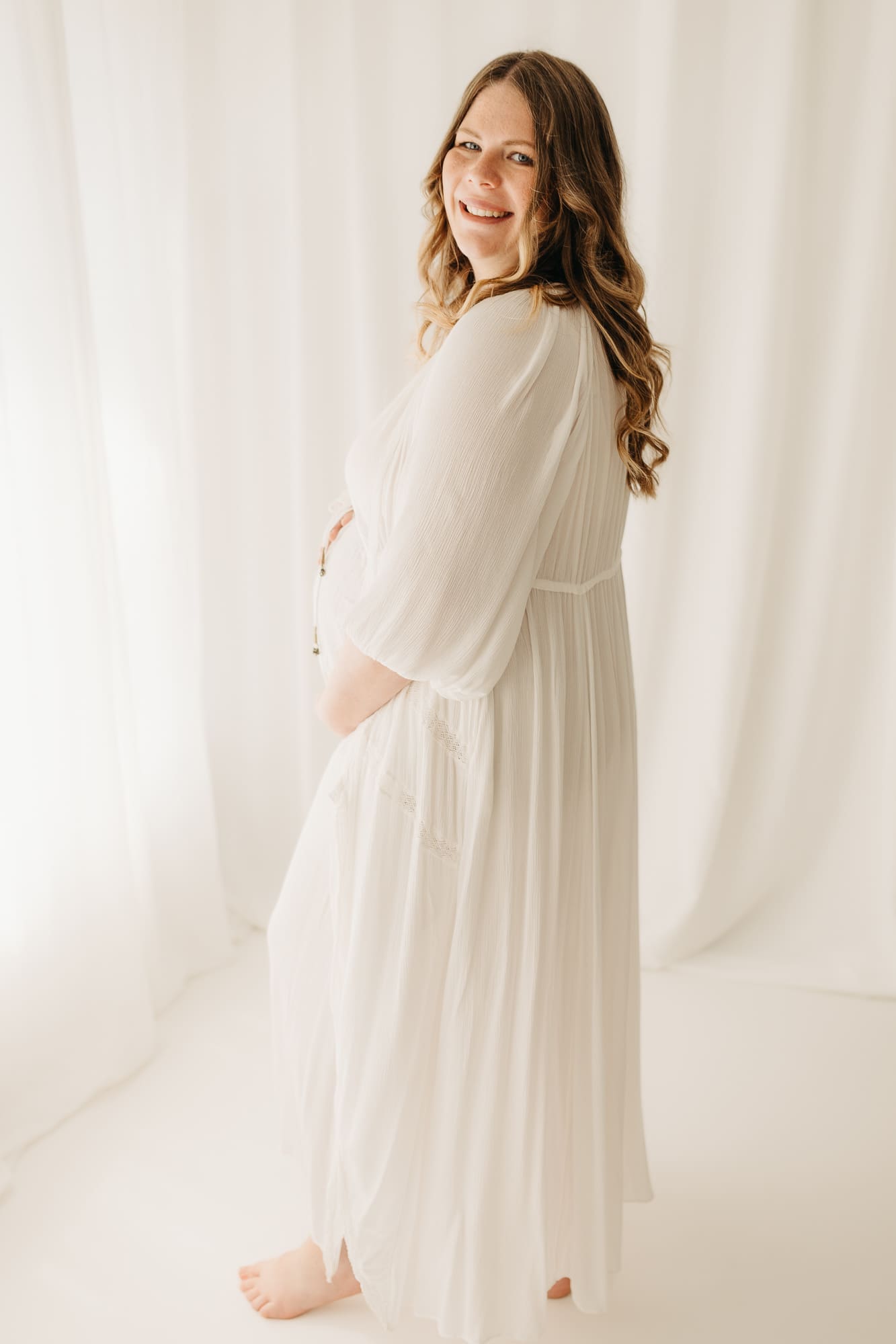 Schwangere Frau im weißen Kleid dreht sich lächelnd um — Babybauch-Shooting im Tageslichtstudio Bottrop